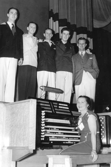 Black and white photo of five people standing on a stage and a person below stage level seated at an organ