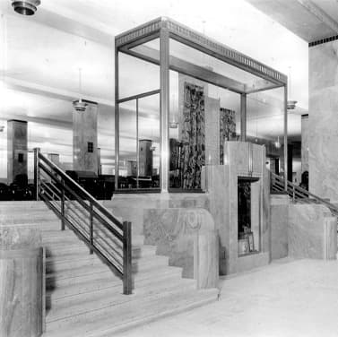 Black and white photo of marble staircases with a mid-century modern design, flanking a glass display case with department store merchandise inside