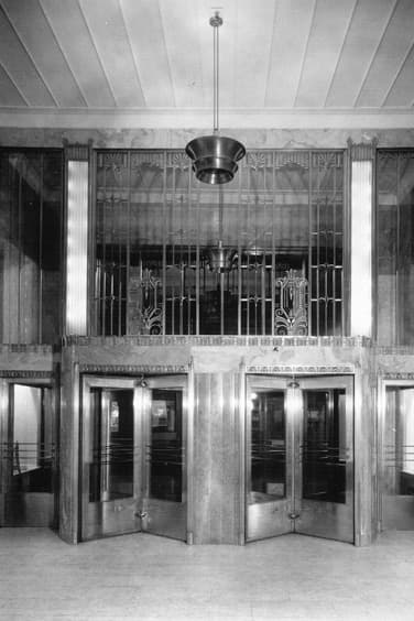 Black and white photo of an art deco lobby with ornate metalwork above a set a double set of revolving doors