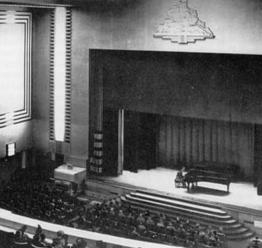 Black and white photo of a concert hall with a grand piano and pianist on stage and a seated audience