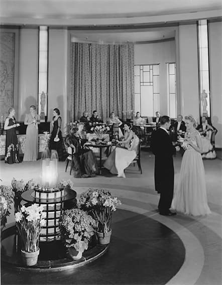 Black and white photo of people in formal attire seated and standing around an art-moderne fountain in a large round dining room
